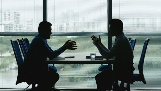 two-human-outlines-against-shuttered-office-window-sitting-opposite-each-other-negotiating